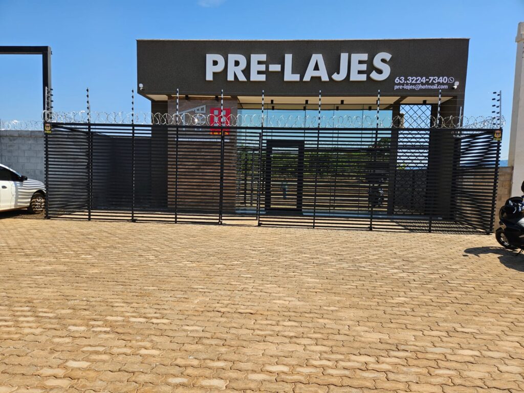 Portão principal da empresa Pré Lajes em Palmas, com estrutura preta, vidro espelhado e cercamento com arame de segurança, vista da rua.
